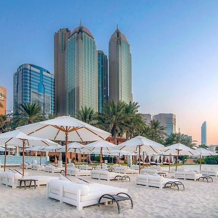 Sheraton Abu Dhabi Hotel&Resort Esterno foto The photo shows a beach scene with white lounge chairs and umbrellas set up on the sand. In the background, there are tall modern buildings, including skyscrapers, suggesting an urban environment near the beach. Palm trees are present, adding a tropi