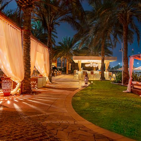 Sheraton Abu Dhabi Hotel&Resort Esterno foto The photo depicts a tranquil outdoor setting at night, featuring a beautifully arranged pathway lined with palm trees. The path is illuminated by soft lights, creating a warm and inviting atmosphere. On one side, there are elegant canopies draped wit