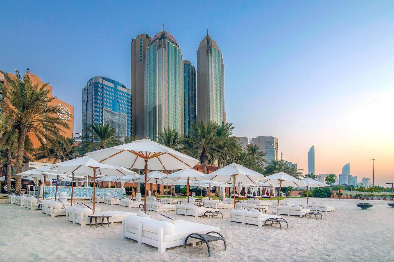 Sheraton Abu Dhabi Hotel&Resort Esterno foto The photo shows a beach scene with white lounge chairs and umbrellas set up on the sand. In the background, there are tall modern buildings, including skyscrapers, suggesting an urban environment near the beach. Palm trees are present, adding a tropi