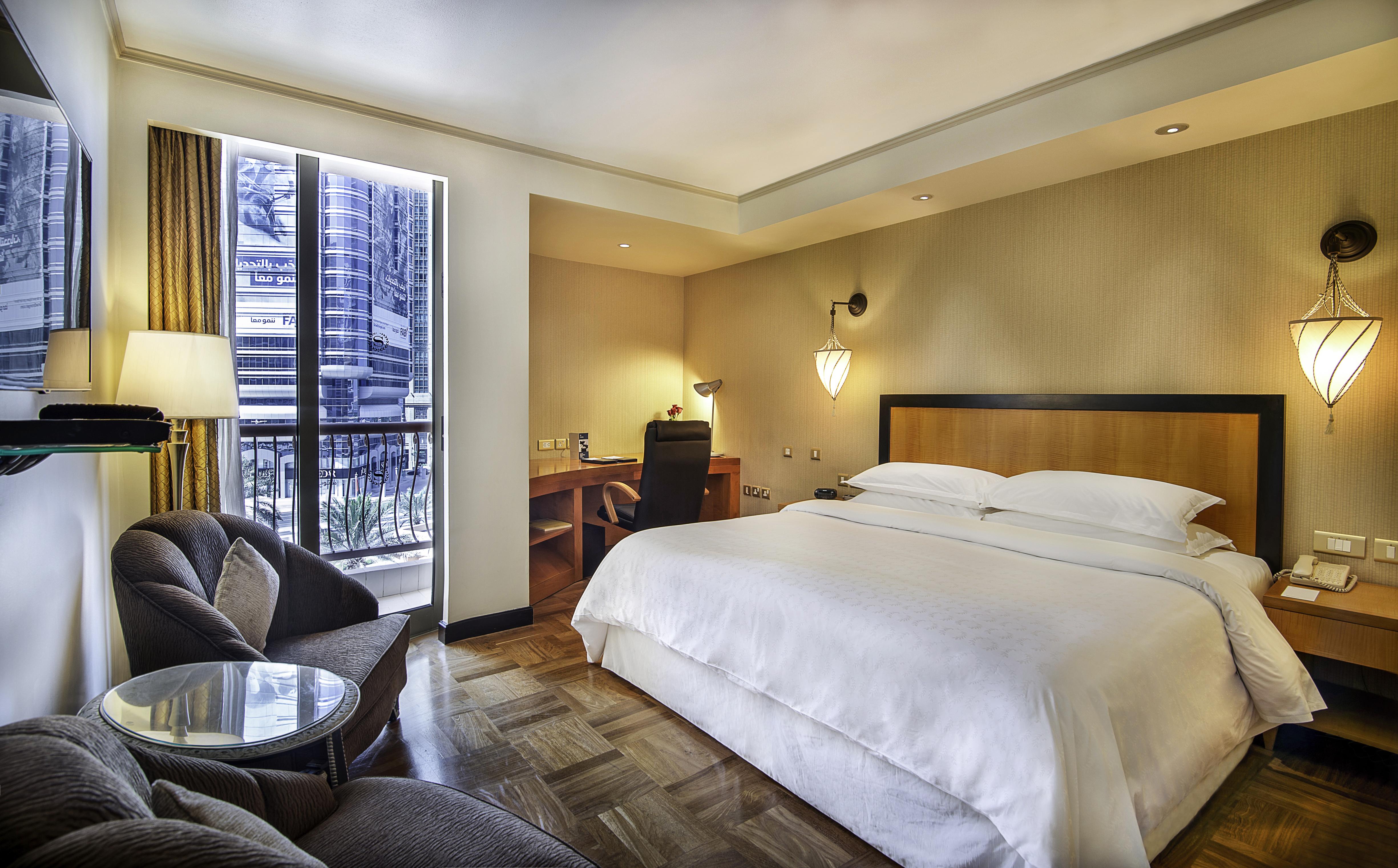 Sheraton Abu Dhabi Hotel&Resort Esterno foto The photo shows a hotel room featuring a large bed with white linens, flanked by bedside tables. There is a comfortable armchair and a small round table in one corner, and a desk with a chair in another area for workspace. Large windows provide natur