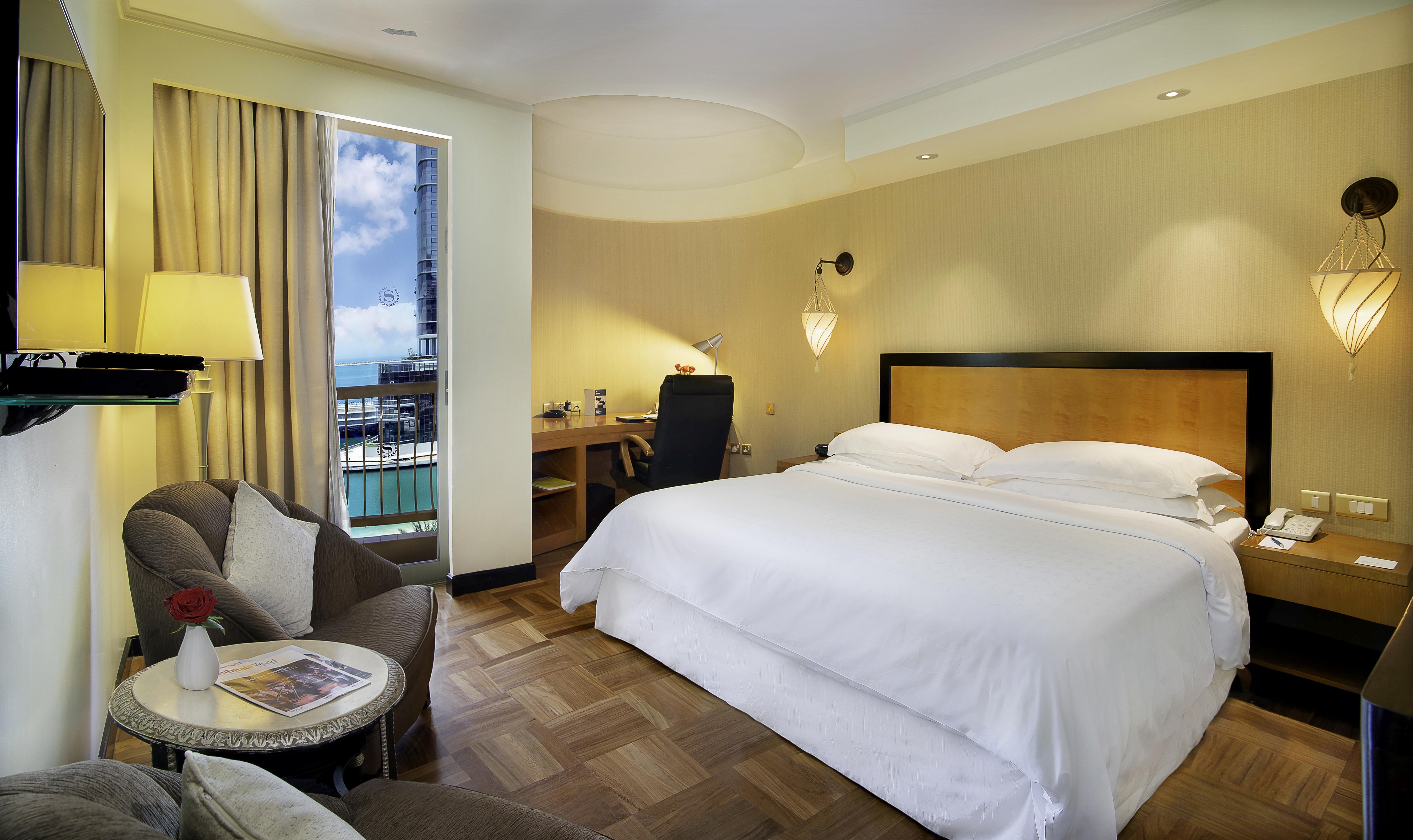 Sheraton Abu Dhabi Hotel&Resort Esterno foto The photo features a modern hotel room. It includes a large bed with white linens, a small seating area with a gray sofa and a round table beside it. There’s a work desk with a black chair, positioned near a window that offers a view outside. The roo
