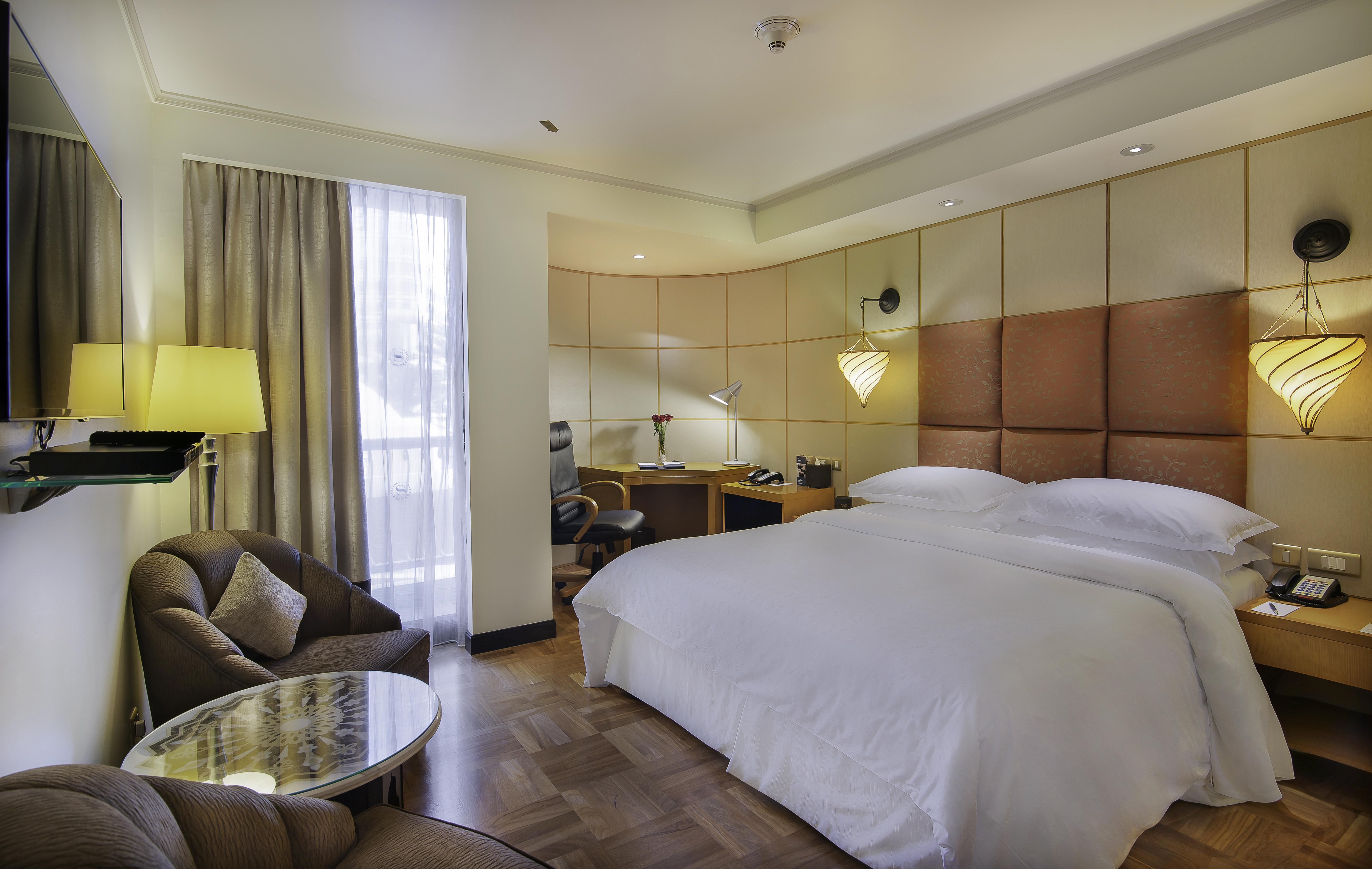 Sheraton Abu Dhabi Hotel&Resort Esterno foto The photo shows a modern hotel room featuring a large bed with white bedding, complemented by a stylish headboard. There is a cozy seating area with a curved brown chair and a small round table. A work desk with a chair is positioned nearby, accompan