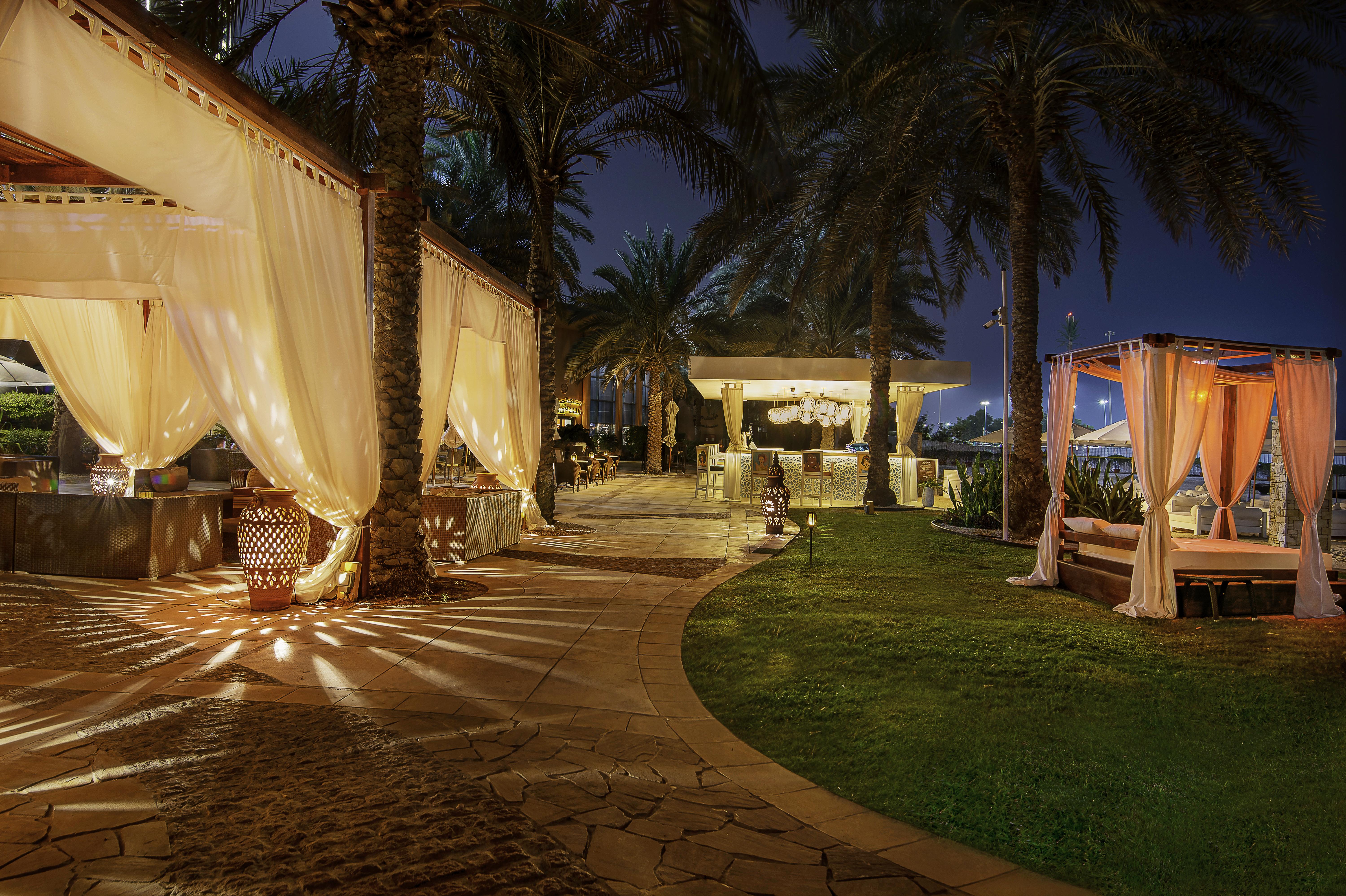 Sheraton Abu Dhabi Hotel&Resort Esterno foto The photo depicts a tranquil outdoor setting at night, featuring a beautifully arranged pathway lined with palm trees. The path is illuminated by soft lights, creating a warm and inviting atmosphere. On one side, there are elegant canopies draped wit