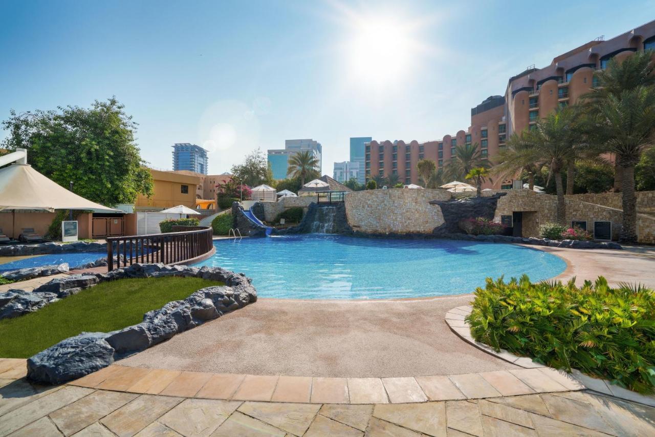 Sheraton Abu Dhabi Hotel&Resort Esterno foto The photo depicts a resort-style pool area. The pool features a large, inviting body of water with a curved design. There are rocky elements along the edges, giving it a natural look. Surrounding the pool are several lounge chairs and shaded cabanas,