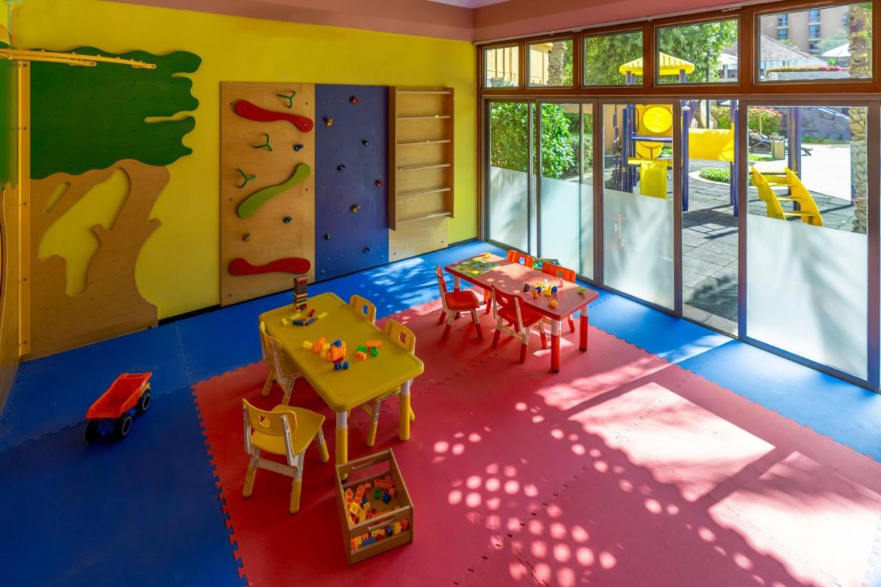 Sheraton Abu Dhabi Hotel&Resort Esterno foto The photo shows a brightly colored children's playroom. The room features a yellow wall with a large green tree design, and another wall with climbing holds for children to engage in physical activity. There are several tables with yellow tabletops a