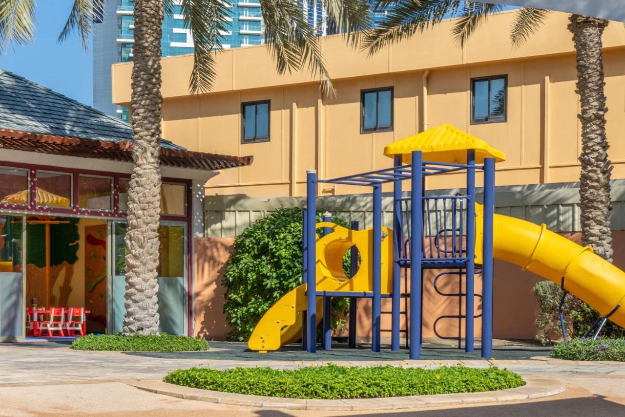 Sheraton Abu Dhabi Hotel&Resort Esterno foto The photo shows a brightly colored playground featuring a yellow slide and climbing structure. The playground is surrounded by lush green landscaping, with palm trees in the background. There is also a building visible to the left, which has large wi
