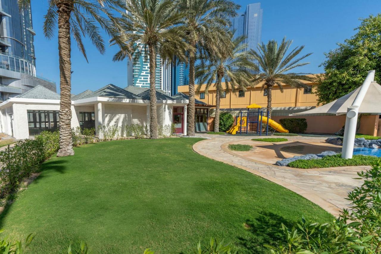 Sheraton Abu Dhabi Hotel&Resort Esterno foto The photo shows a landscaped outdoor area featuring a lush green lawn and several palm trees. In the background, there are modern buildings, suggesting an urban environment. There is a playground with a yellow slide visible, indicating a children's p