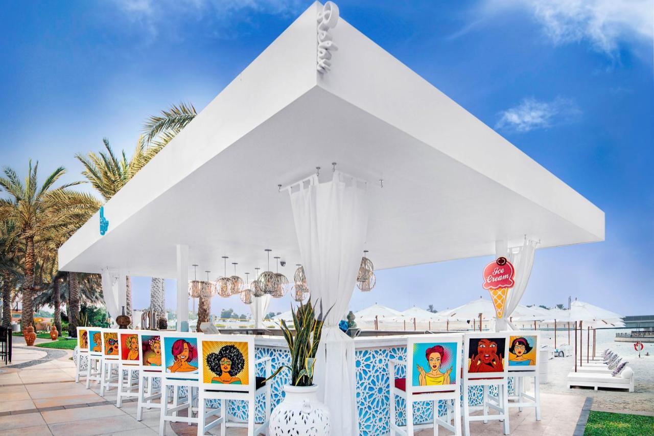Sheraton Abu Dhabi Hotel&Resort Esterno foto The photo shows a stylish outdoor bar area with a large white canopy. The bar has colorful paintings on the backs of the white chairs, depicting various artistic representations, likely of women. There are decorative elements such as hanging lanterns