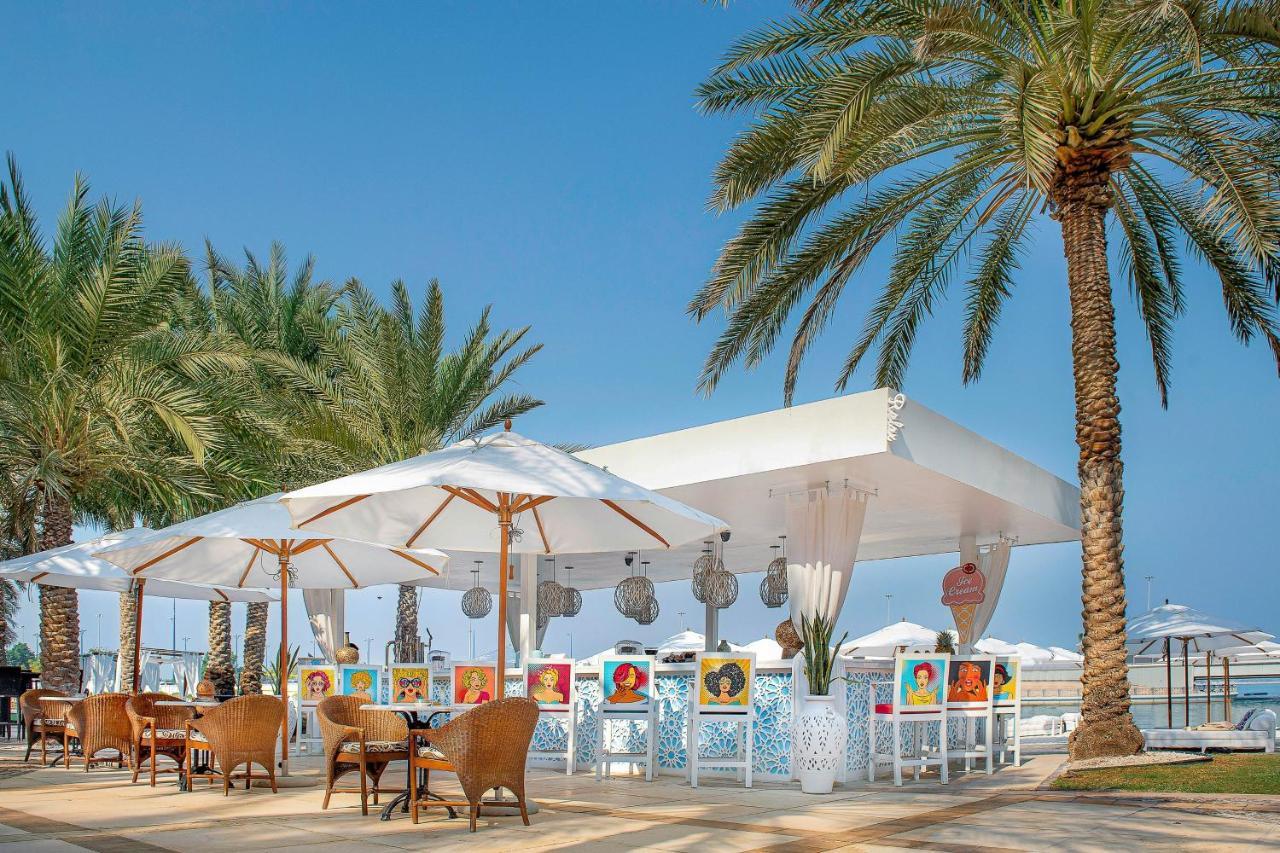 Sheraton Abu Dhabi Hotel&Resort Esterno foto The photo shows an outdoor café or restaurant setup near a beach or waterfront. It features a white canopy providing shade over several tables. There are stylish wicker chairs arranged around these tables, and the setting is decorated with colorful a
