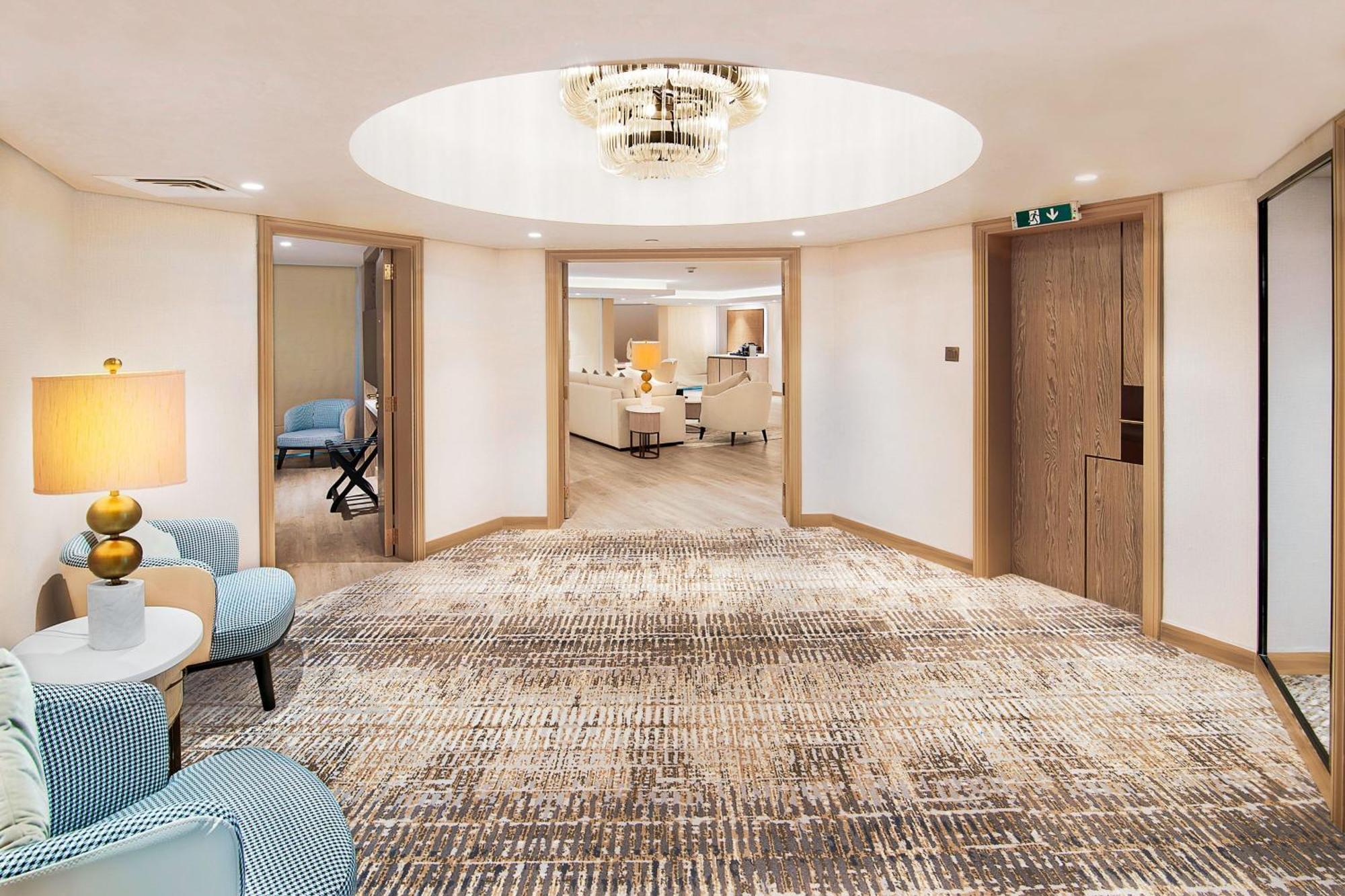 Sheraton Abu Dhabi Hotel&Resort Esterno foto The photo shows a modern, well-lit hallway with a circular ceiling feature that includes a decorative light fixture. The flooring is covered with a textured carpet that has a subtle pattern. On the left, there are two light blue chairs with a round t