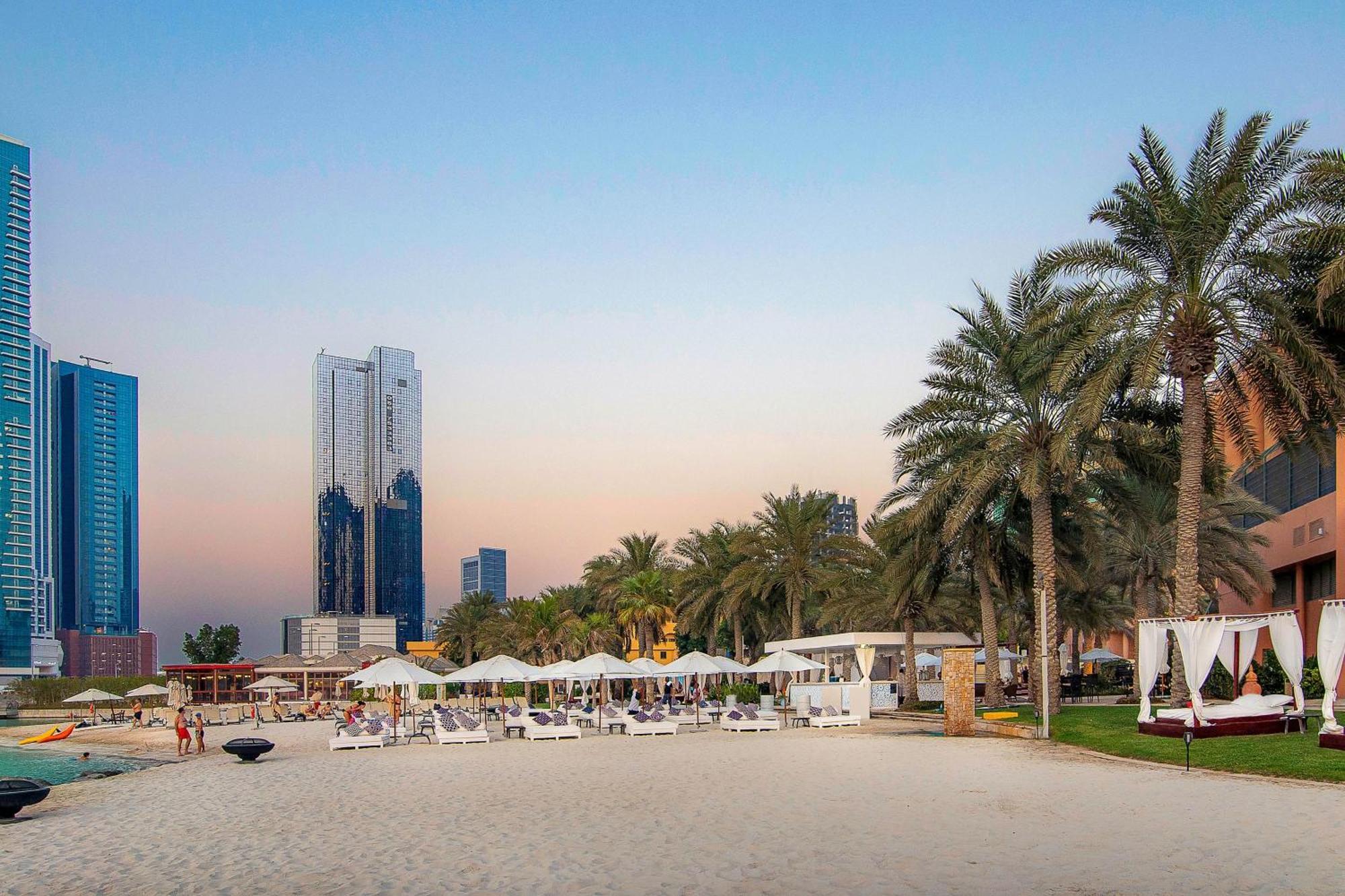 Sheraton Abu Dhabi Hotel&Resort Esterno foto The photo depicts a beach scene featuring a sandy shoreline with palm trees lining the area. There are several lounge chairs and shaded cabanas set up for relaxation, suggesting a resort-like atmosphere. In the background, modern skyscrapers rise, sh