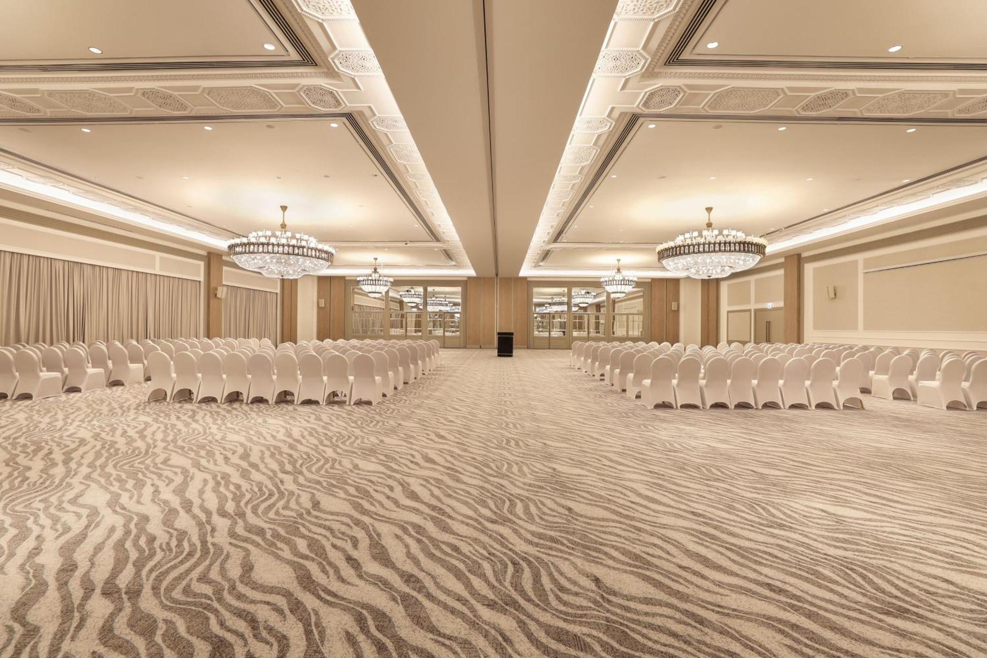 Sheraton Abu Dhabi Hotel&Resort Esterno foto The photo shows an elegant indoor venue, likely set up for an event or conference. The room features rows of white chairs arranged facing a central area, with a light-colored, patterned carpet resembling zebra stripes. It is illuminated by stylish ch