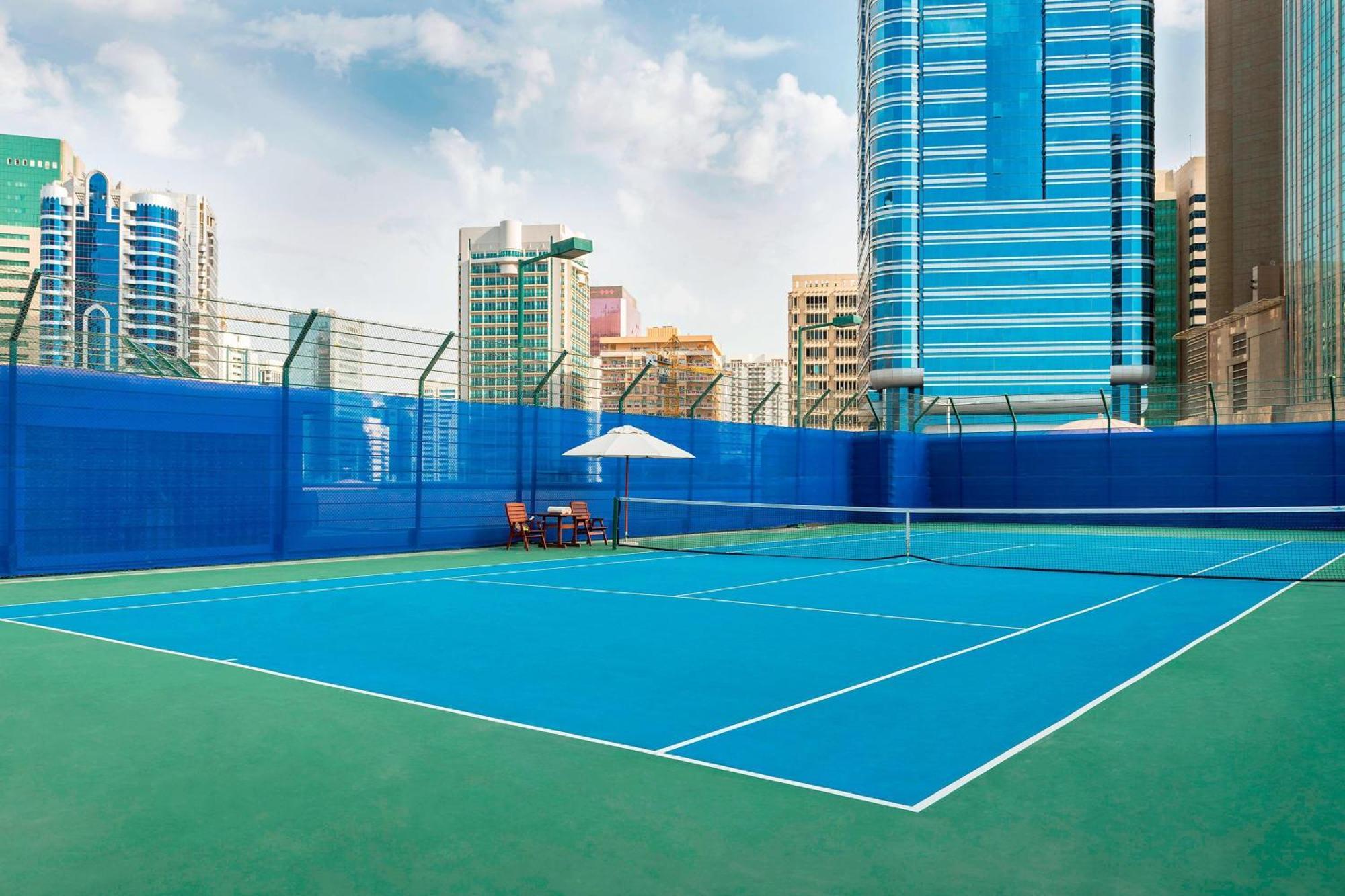Sheraton Abu Dhabi Hotel&Resort Esterno foto The photo shows a tennis court situated in an urban environment with tall buildings in the background. The court features a vibrant blue playing surface, surrounded by a green area. There is a net in the center, and a couple of chairs and an umbrella