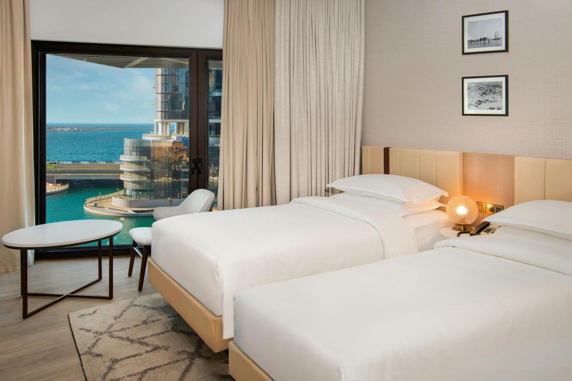 Sheraton Abu Dhabi Hotel&Resort Esterno foto The photo shows a modern hotel room featuring two twin beds with white bedding. The room has a neutral color palette, with curtains covering large windows that offer a view of the ocean and a cityscape outside. A small round table and a chair are pos