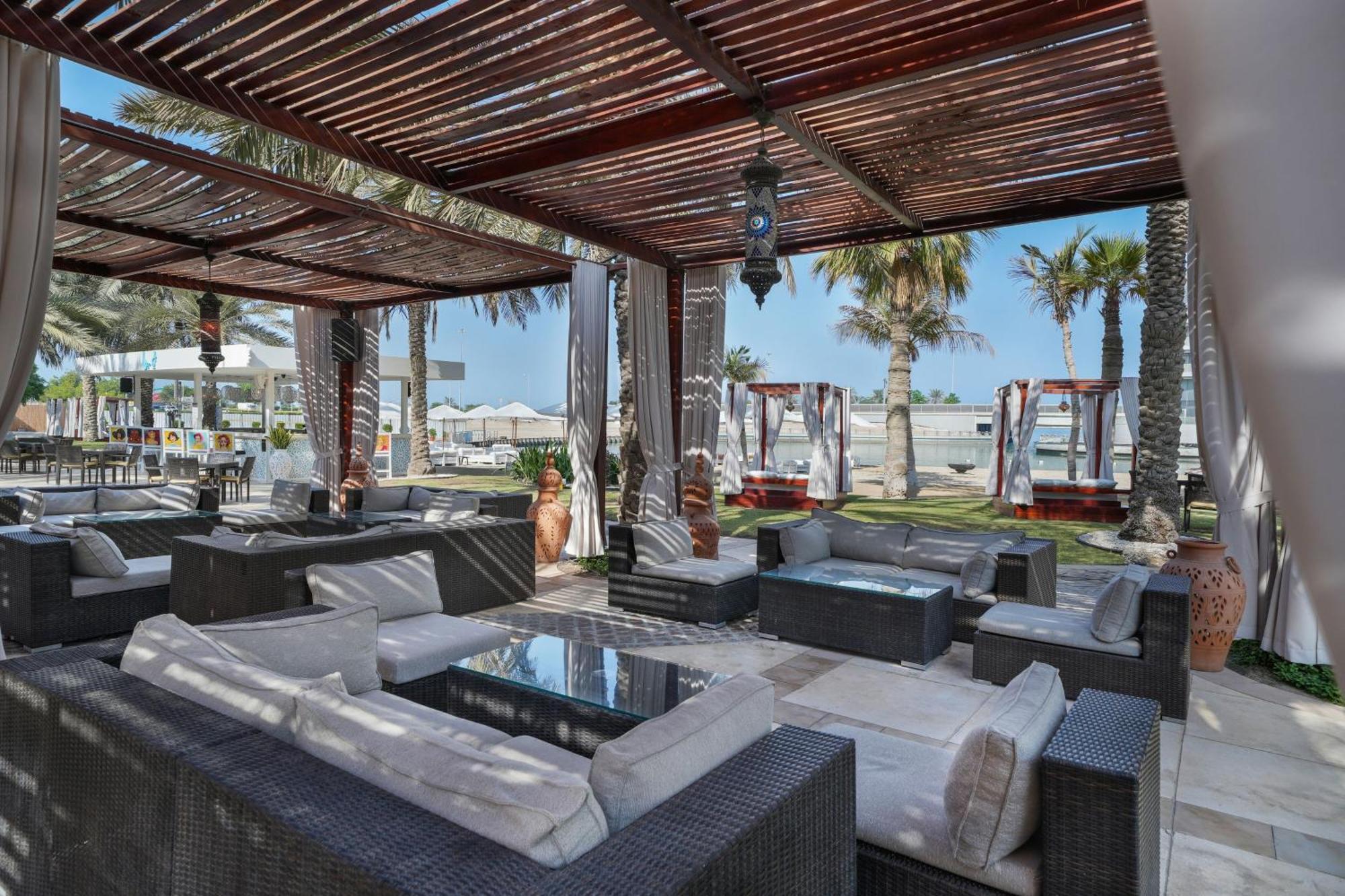 Sheraton Abu Dhabi Hotel&Resort Esterno foto The photo shows a stylish outdoor lounge area, likely at a beach resort. It features comfortable seating made of dark wicker furniture with plush cushions. The area is shaded by a wooden pergola, creating a cozy atmosphere. In the background, there a