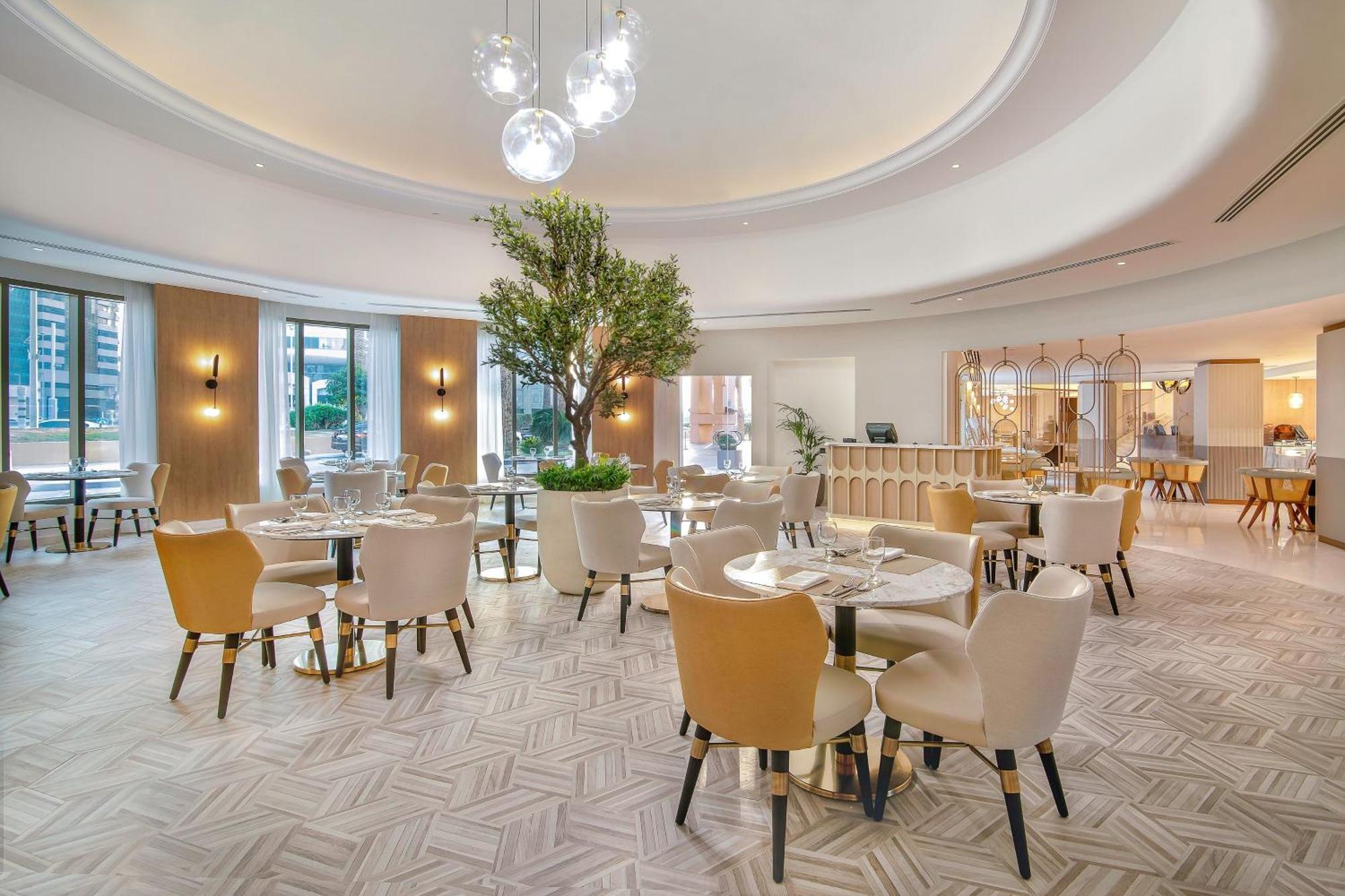 Sheraton Abu Dhabi Hotel&Resort Esterno foto The photo shows a modern restaurant interior. It features elegantly arranged tables with beige and light-colored chairs. The decor includes a large central tree, adding a touch of greenery, and stylish lighting fixtures hanging from the ceiling. The 