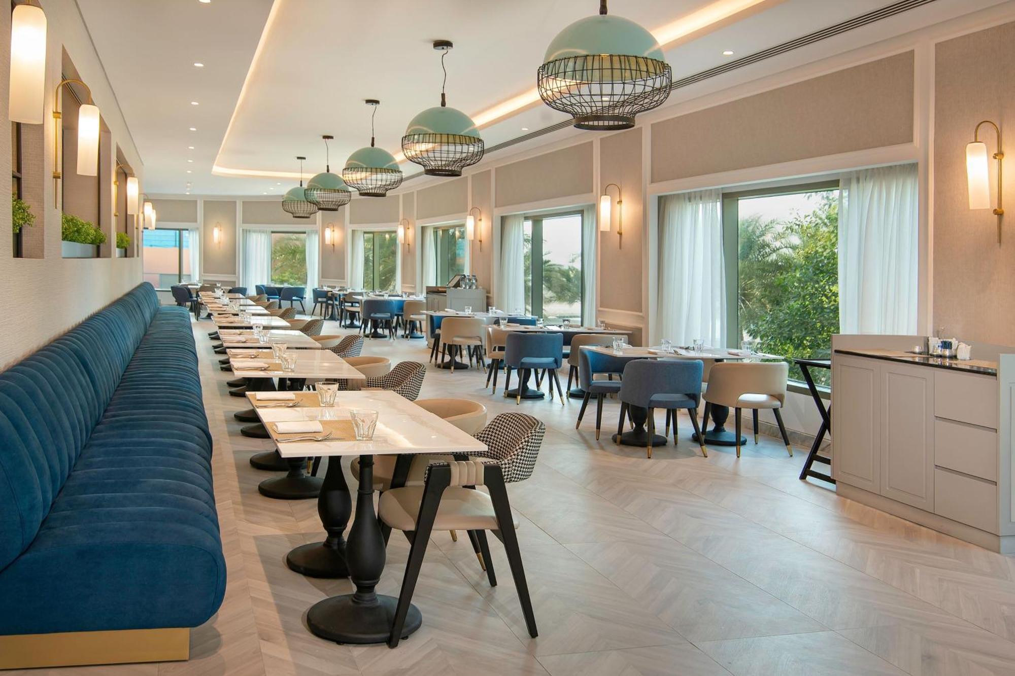Sheraton Abu Dhabi Hotel&Resort Esterno foto The photo depicts a modern restaurant interior with a spacious and inviting atmosphere. There are several tables arranged neatly throughout the dining area, each set with white plates and cutlery. Long blue banquettes line one side of the space, prov
