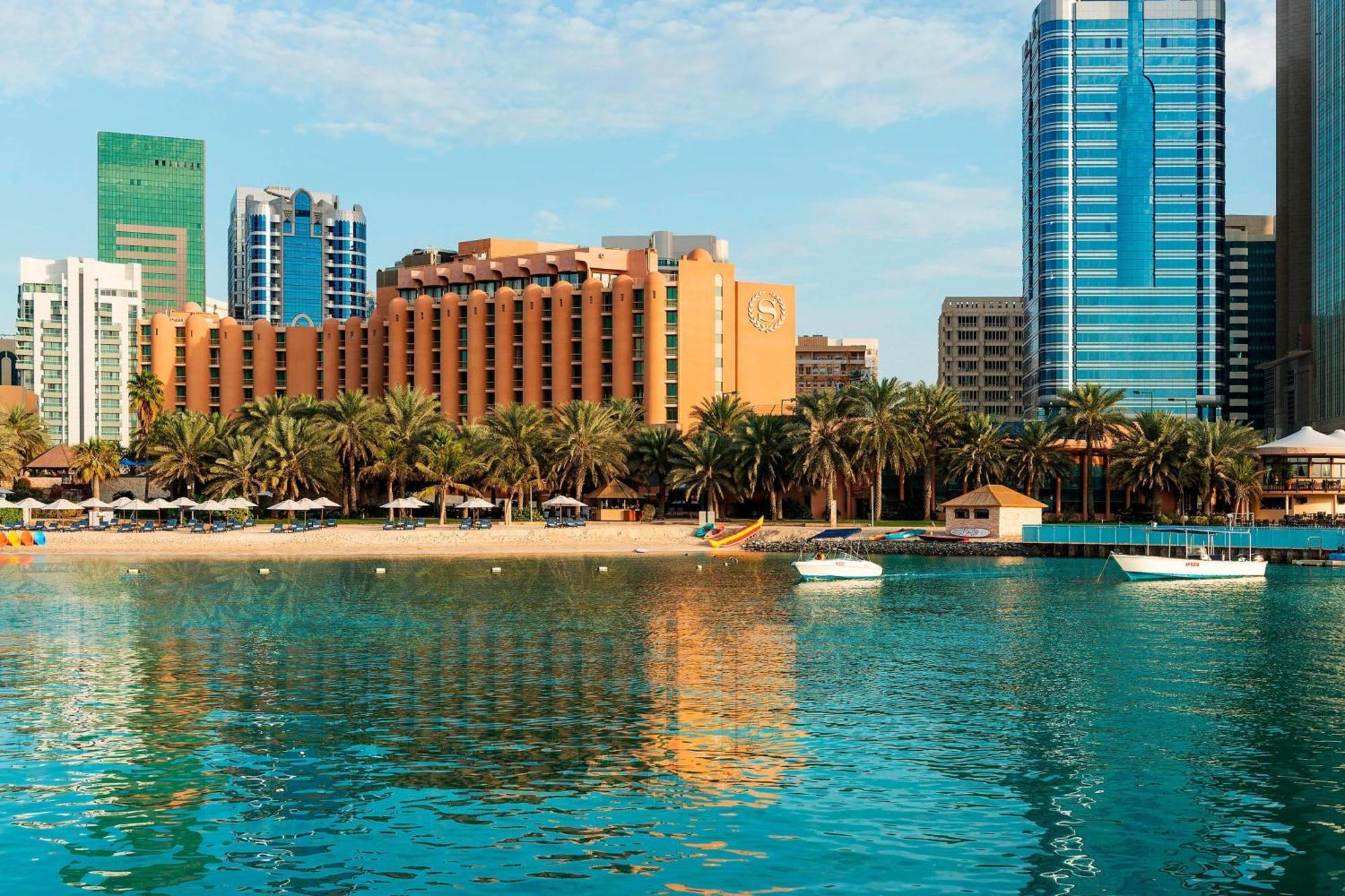 Sheraton Abu Dhabi Hotel&Resort Esterno foto The image depicts a coastal scene with a clear blue sky reflecting on the water. In the foreground, there is a beach with palm trees and small boats moored in the water. The backdrop features a mix of modern buildings, including a large hotel and hig