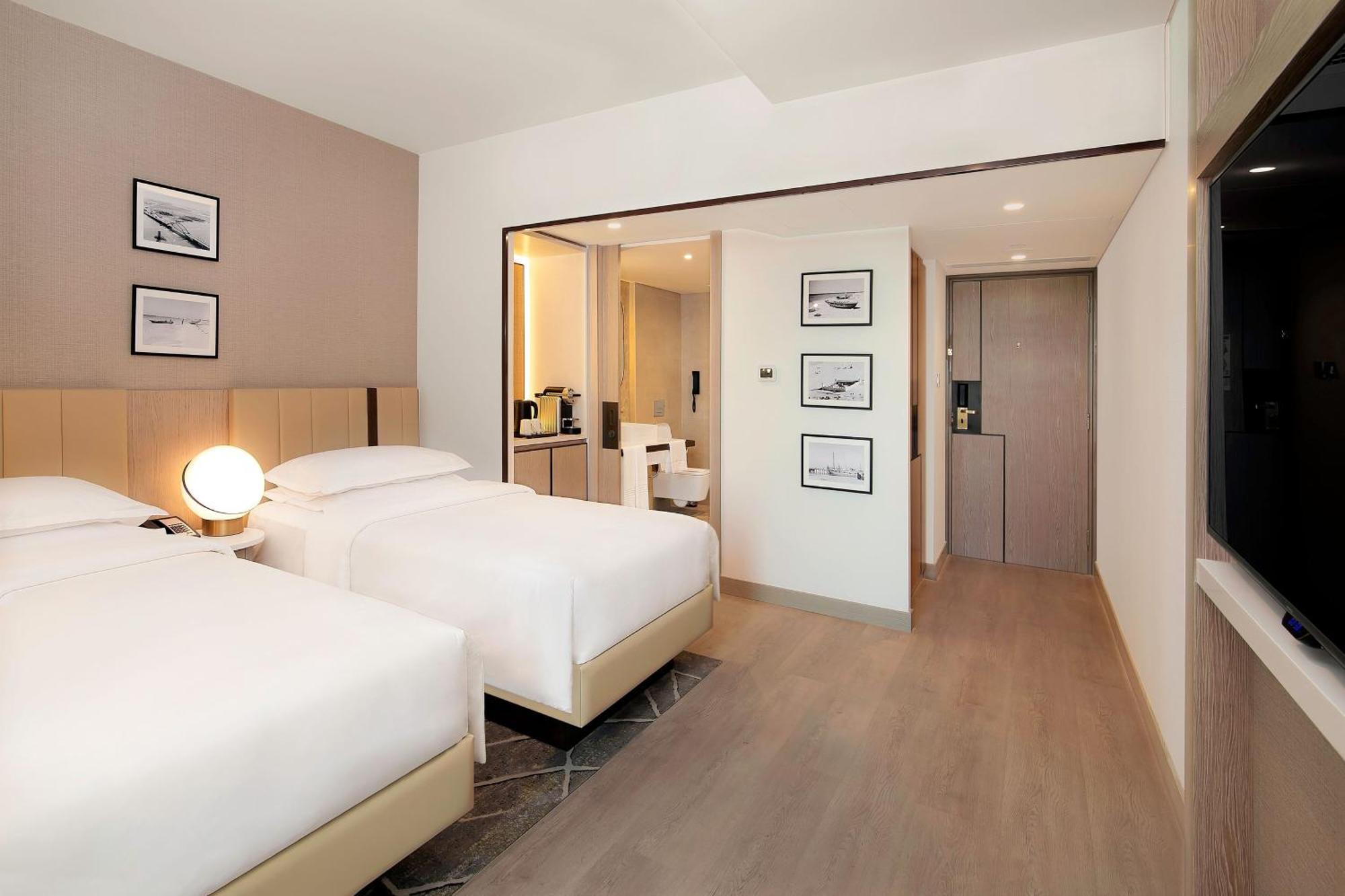 Sheraton Abu Dhabi Hotel&Resort Esterno foto The photo shows a modern hotel room featuring two single beds with white bed linens. The room has light-colored walls and wooden flooring. There are framed black-and-white photographs on the wall above the beds. A round lamp is placed on a nightstand